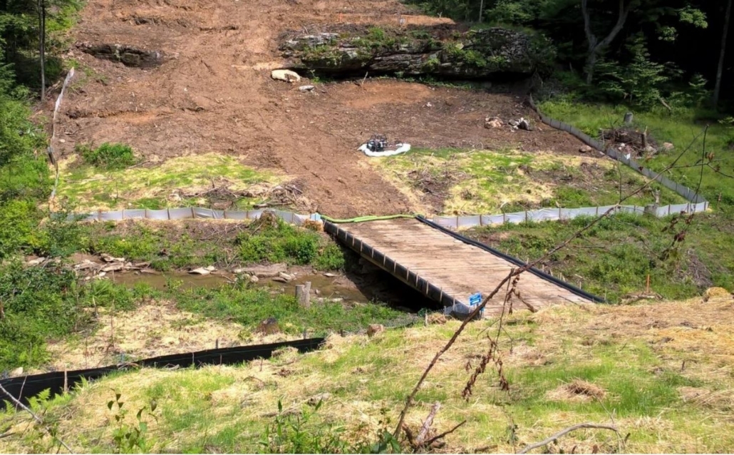 Image of a stream being crossed by MVP by way of a timber mat bridge. The mountainside is torn through, and a silt fence blocks the stream from runoff and contamination.