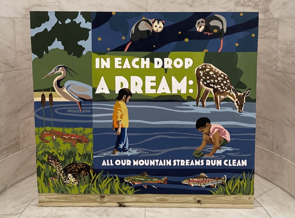 Image of the mural in the WV State Capitol Rotunda. The mural is framed the white marble walls and in bold white letters, the mural reads, "In each drop a dream: All our mountain streams run clean." The mural depicts WV Stream Life and the magic within it. A Great Blue Heron stands in the water while a white tail deer takes a drink. A newt and turtle hide in the grass with opossums on a branch over head. In the creek, two children play, and one, with wonder in their eyes, scoops a crawdad from the creek onto a petal. Native Brook Trout and Rainbow Trout swim below the surface.
